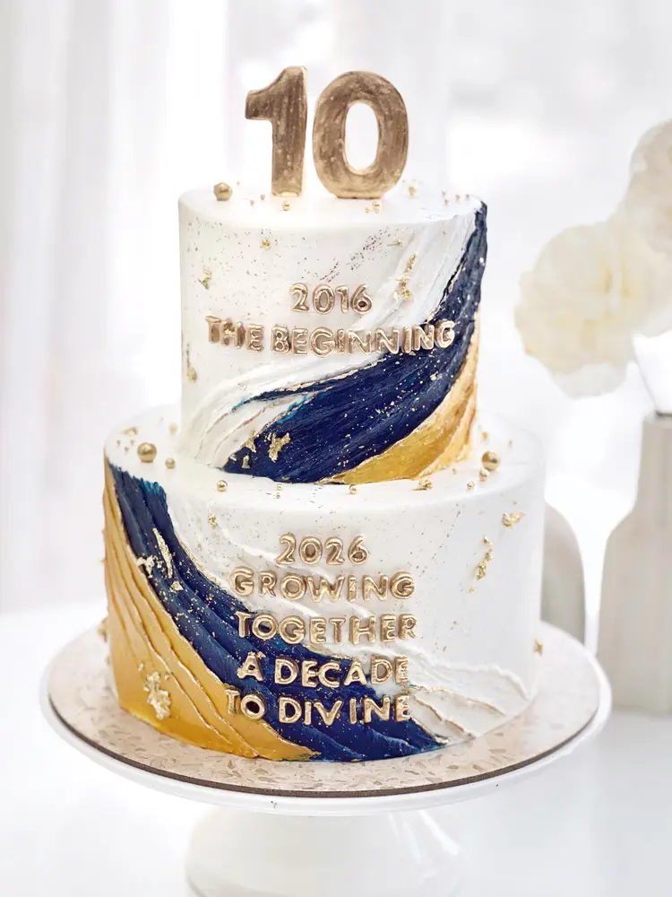 Cake dua tingkat putih dengan sapuan biru navy dan emas, angka 10 di atasnya