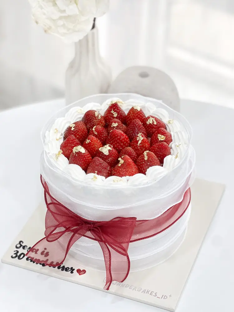 Cake putih dengan strawberry segar di atasnya dan pita merah