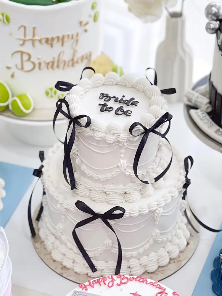 Cake dua tingkat putih bergaya vintage dengan pita hitam, bertuliskan Bride to be